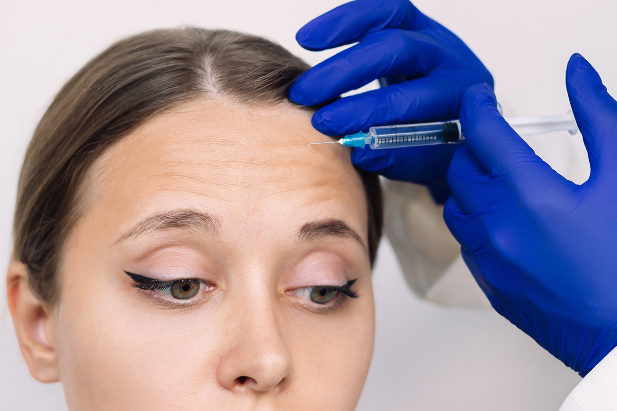 Young woman and doctor s hands in blue gloves holding syringe needle for injection of facial wrinkles on the forehead